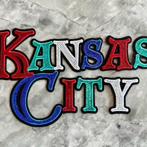 May include: A colorful patch with the words "Kansas City" in a glittery, multi-colored font. The letters are outlined in black and have a 3D effect.