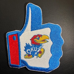 May include: Blue and white embroidered patch with a thumbs up hand shape. The hand is holding a cartoon mascot of a blue bird with a red head and yellow shoes. The bird is running and has the letters "KU" on its chest.