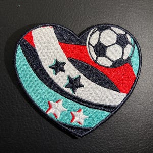 May include: A heart-shaped embroidered patch with a soccer ball, stars, and red, white, and blue stripes on a teal background.