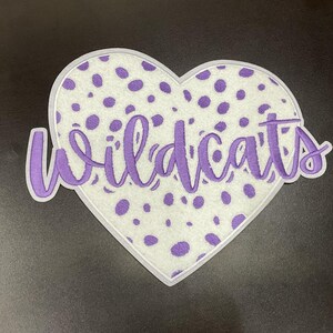 May include: A heart-shaped patch with a white background and purple polka dots. The word "Wildcats" is written in purple script across the heart. The patch has a white border.