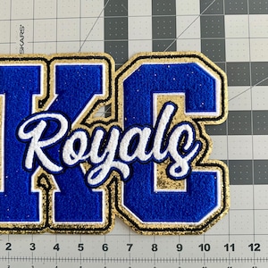 May include: A blue and gold embroidered patch with the letters "KC" and the word "Royals" in white script. The patch has a gold glitter border with a black outline. The patch is on a gray grid background.