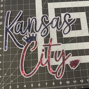 May include: A white fabric patch with the words "Kansas City" in blue and red sequins. The word "Kansas" is in blue sequins and the word "City" is in red sequins. A crown is above the word "Kansas".