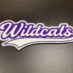 May include: A purple and white Wildcats patch with a white border. The word "Wildcats" is embroidered in a script font with purple thread and a white outline. The patch has a white banner shape at the bottom.