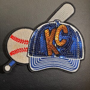 May include: A blue and white baseball cap with a gold and orange sequined "KC" on the front. A baseball and a bat are attached to the side of the cap.