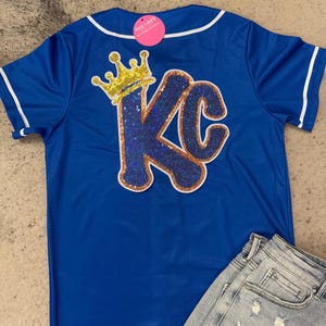 Op de afbeelding: Een koninklijk blauw baseballshirt met een witte kraag en witte afwerking rond de mouwen. De achterkant van het shirt heeft een grote, glinsterende gouden en blauwe "KC" met een gouden kroon erboven.
