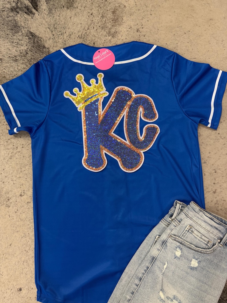 Op de afbeelding: Een koninklijk blauw baseballshirt met een witte kraag en witte afwerking rond de mouwen. De achterkant van het shirt heeft een grote, glinsterende gouden en blauwe "KC" met een gouden kroon erboven.