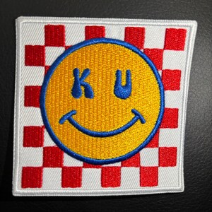 May include: A white and red checkered patch with a yellow smiley face with blue eyes and a blue mouth. The smiley face has the letters "KU" inside of it.