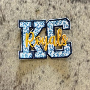 May include: Blue and white embroidered patch with the letters "KC" in a varsity style font. The word "Royals" is embroidered in yellow below the letters.