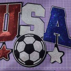 May include: A set of sequined patches featuring the letters U, S, and A in red, white, and blue. The letter U is red, S is white, and A is blue. Below are a black and white soccer ball patch, a red star patch, and a white star patch.