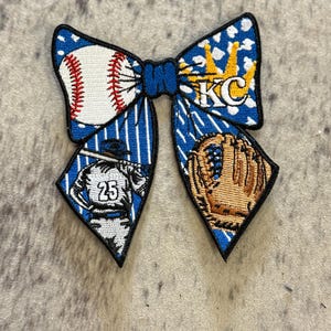 May include: A blue and white baseball-themed bow with a baseball, glove, and the letters "KC". The bow has a black border and is decorated with baseball-related imagery. The bow is a decorative accessory.