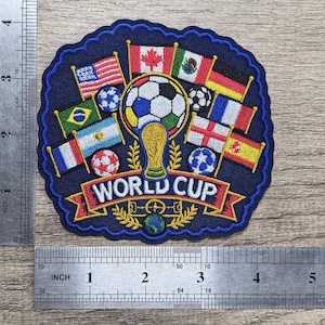 May include: A circular embroidered patch featuring a soccer ball, a gold trophy, and various national flags. The text WORLD CUP is centered on a red banner. A metal ruler shows the patch is approximately 3.5 inches wide and 3.5 inches tall.