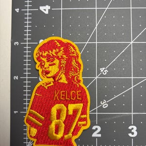 May include: Embroidered patch featuring a stylized portrait of a person in a red and gold jersey. The jersey displays the name "KELCE" and the number "87" in gold. The patch has a yellow border and is likely for apparel.