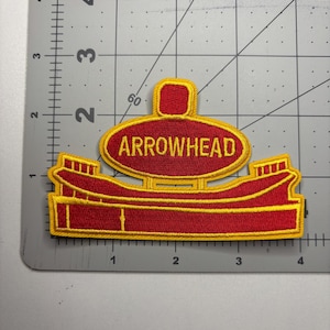 May include: Embroidered patch in red and gold, featuring the word "ARROWHEAD" in a gold oval. The patch has a unique shape with a rectangular top and a stylized, boat-like bottom. The patch is approximately 4 inches wide.