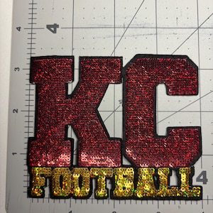 May include: A red sequined patch with the letters "KC" above the word "FOOTBALL" in gold sequins. The letters are outlined in black. The patch is on a gray surface with measurement markings.