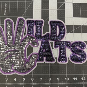 May include: Purple and silver sequined iron-on patch with the words "Wildcats" and a hand with three fingers raised.