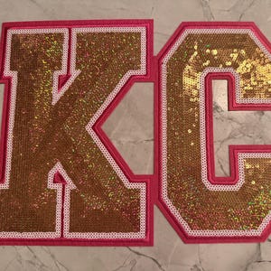 May include: Two large, embroidered patches spelling out "KC". The letters are gold sequins with a white border, set against a hot pink background. The patches are likely for a jacket or bag.