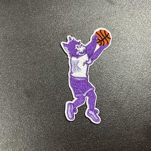 May include: A purple embroidered patch featuring a cartoon wolf in a basketball uniform, holding an orange basketball. The wolf is wearing a white jersey with a purple "K" on it, purple shorts, and purple shoes. The patch has a white border.