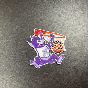 May include: A purple and white patch featuring a cartoon cat dunking a basketball. The cat is wearing a jersey with the letter "K". The basketball hoop is orange and red, and the ball is orange.