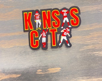 May include: A red and yellow embroidered patch with the words "Kansas City" and four football players in red and white uniforms.