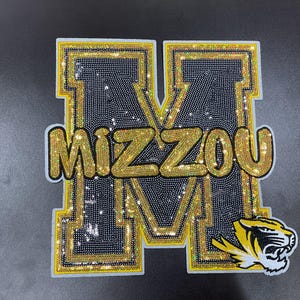 May include: A large, decorative patch featuring the word "MIZZOU" in gold, sequined letters over a black sequined background. The letter "M" is outlined in gold and has a yellow and black tiger head logo on the lower right.