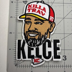 May include: Embroidered patch featuring a cartoon image of a man wearing a white and red baseball cap with the text "Killa Trav" and the number 87. The patch also includes the name "KELCE" in bold black letters and a small "KC" logo.