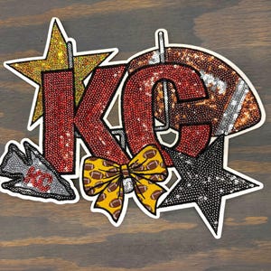 May include: A red, white, and gold sequined applique featuring the letters "KC" with a football helmet, a star, a bow, and an arrow with the letters "KC".
