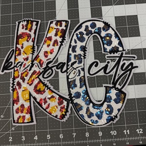 May include: A white patch with the letters "KC" in large, colorful, sequined letters. The "K" is red, yellow, and white with a leopard print pattern. The "C" is blue, white, and gold with a leopard print pattern. The letters are outlined in black and the words "Kansas City" are written in black cursive script below the letters.