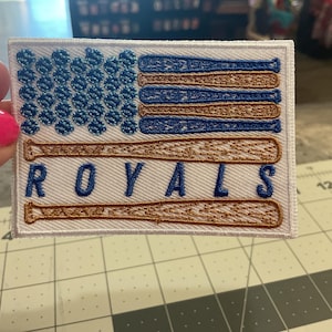May include: Embroidered patch featuring a design inspired by the American flag, with blue baseballs in the upper left corner and baseball bats in place of stripes. The word "ROYALS" is embroidered in blue.