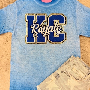 May include: A light blue t-shirt with a blue and gold glitter "KC Royals" patch on the front. The shirt is a faded ombre style.