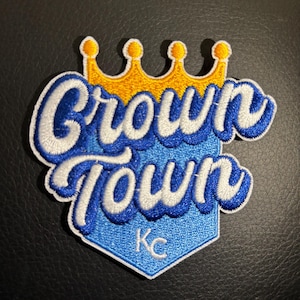 May include: A blue and white embroidered patch with the words "Crown Town" in a stylized font. A yellow crown sits above the words. The patch has a blue diamond shape with the letters "KC" in white in the center.