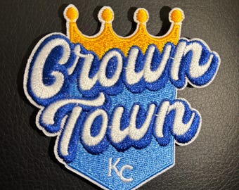 May include: A blue and white embroidered patch with the words "Crown Town" in a stylized font. A yellow crown sits above the words. The patch has a blue diamond shape with the letters "KC" in white in the center.