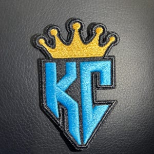 May include: A blue and black embroidered patch with the letters "KC" in a stylized font. A gold crown sits above the letters.