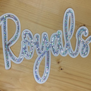 May include: A decorative sign with the word "Royals" in a cursive font. The letters are filled with iridescent sequins and outlined in white. The sign is on a light brown wooden surface.