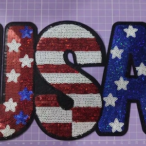 May include: A set of three large letters spelling USA, crafted with red, white, and blue sequins. The letter U features a red background with white and blue stars, the letter S has red and white horizontal stripes, and the letter A is blue with white stars.