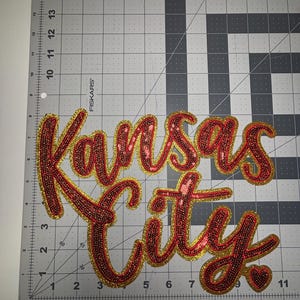 May include: Red sequined patch with the words "Kansas City" in a cursive font. The letters are outlined in gold glitter. The patch is on a gray cutting mat with measurement markings.