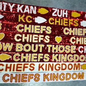 May include: A collection of beaded ribbons in red, white, and gold, featuring text related to the Kansas City Chiefs. The ribbons display phrases like "CITY KAN", "CHIEFS", and "KINGDOM", along with arrow and heart symbols.