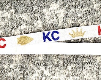May include: White fabric strap with gold crowns and the letters "KC" in red, blue, and gold.