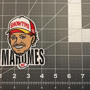 May include: Embroidered patch featuring a cartoon portrait of a smiling man wearing a red, white, and yellow baseball cap with the word "SHOWTIME". Below the portrait is the name "MAHOMES" in bold black letters, with a "KC" logo.