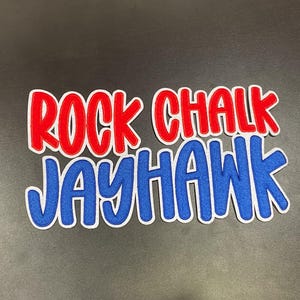 May include: A die-cut sticker with the words "ROCK CHALK JAYHAWK" in red and blue, with a white outline. The sticker is on a dark gray background.