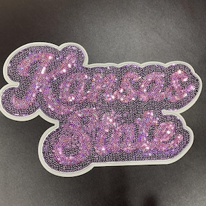 May include: A purple sequined patch with the words "Kansas State" in a cursive font. The patch has a white border and is on a dark background. The sequins are a mix of light and dark purple.
