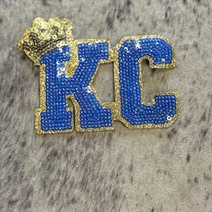 May include: A blue and gold sequined patch with the letters "KC" and a gold crown above the letters.