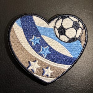 May include: A blue, white, and brown heart-shaped patch with a soccer ball and stars. The soccer ball is black and white and is partially visible in the upper right corner of the patch.