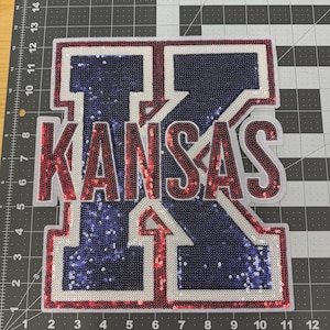 May include: A sequined patch featuring the word "KANSAS" in red sequins, outlined in white, over a large blue sequined "K" with a white outline. The patch is rectangular with a white border. The number "1" is visible.