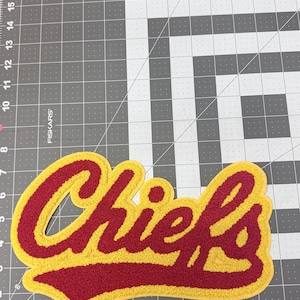 May include: A red and yellow Chiefs patch with a textured, embroidered appearance. The word "Chiefs" is in a stylized font with a yellow outline and red fill. The patch is on a gray cutting mat.
