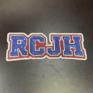 May include: A rectangular patch with the letters "RCJH" in a collegiate font. The letters are blue with red outlines and filled with sparkling rhinestones. The patch has a white border and is set against a dark gray background.