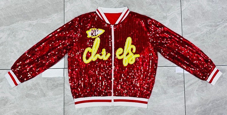 K&ouml;nnte beinhalten: Rote Pailletten-Bomberjacke mit wei&szlig;-roten Streifen. Mit dem Text "Chiefs" in gelber Schrift und einem "KC"-Logo. Die Jacke hat einen durchgehenden Rei&szlig;verschluss und einen wei&szlig;en Kragen.