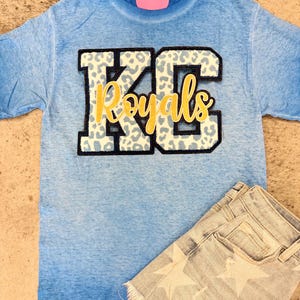 May include: A blue tie-dye t-shirt with a white and black leopard print applique of the letters "KC" and the word "Royals" in gold lettering.