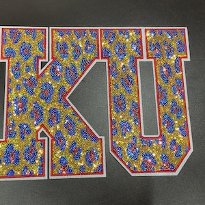 May include: Embroidered patches spelling "KU" in a leopard print design. The letters are gold with blue and red accents, outlined in red, and bordered with white. The patches are likely for apparel.