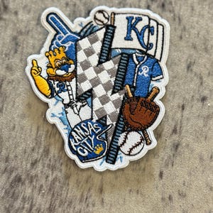 May include: A white embroidered patch with a blue and white checkered lightning bolt, a baseball, a baseball bat, a baseball glove, a baseball jersey, a baseball cap, and a cartoon mascot. The patch says "Kansas City" and "KC".