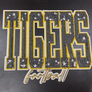 May include: A decorative patch with the word "TIGERS" in large, black, and yellow letters, embellished with small, sparkling accents. The word "football" is written below in a cursive font.